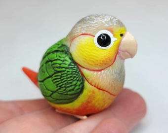 May include: A small, hand-painted ceramic figurine of a green and yellow parakeet. The bird has a red tail and a white head with black eyes.
