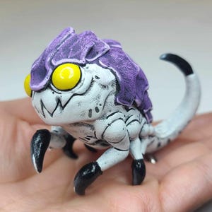 Puede incluir: Una pequeña criatura blanca pintada a mano con detalles en morado y negro. La criatura tiene una cola larga, cuatro patas y una cabeza grande con ojos amarillos.