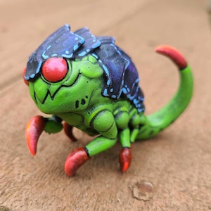 May include: A green and blue hand-painted creature with red eyes and a long tail. The creature has a segmented body and appears to be made of clay.