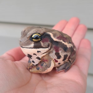May include: A hand-painted ceramic toad figurine with brown, black, and red markings. The toad has a yellow eye with a black pupil.