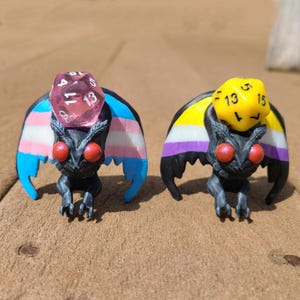 May include: Two black moth figurines with wings painted in the colors of the transgender and genderqueer pride flags. Each moth is wearing a clear plastic dice with numbers on the sides.