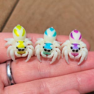 Könnte beinhalten: Drei weiße Springspinnen-Figuren mit bunten Akzenten. Die Spinnen haben gelbe, blaue und violette Akzente auf ihren Körpern.