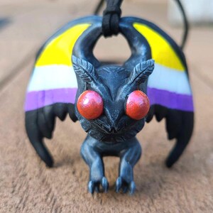 May include: A handcrafted pendant featuring a stylized moth with large red eyes and wings painted in the non-binary pride flag colors: yellow, white, purple, and black. The moth is suspended from a black cord, perfect for wearing as a necklace.