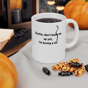 Può includere: Tazza in ceramica bianca con liquido scuro, con la scritta "Scottie, don't beat me up yet, I'm having a shi". La tazza è su una superficie grigia, con caramelle di Halloween e una zucca sullo sfondo.