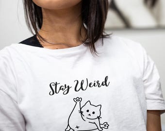 May include: White t-shirt with black text that says "Stay Weird" and a cartoon cat illustration.