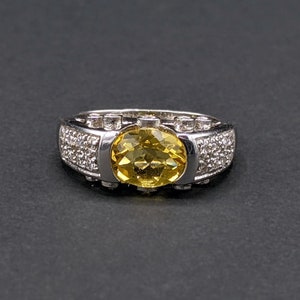 Op de afbeelding: Een zilveren ring met een gele edelsteen omringd door kleinere heldere edelstenen.