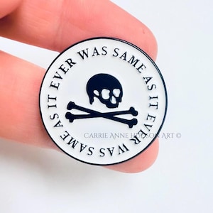 May include: A round, white enamel pin with a black skull and crossbones design. The text "AS IT EVER WAS SAME AS IT EVER WAS" is printed around the edge in black. The pin is held between fingers.