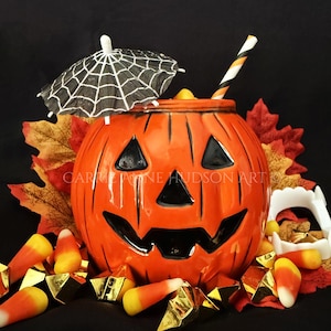 May include: A bright orange ceramic jack-o'-lantern mug with a carved face, featuring a black and white spiderweb cocktail umbrella, a striped straw, scattered candy corn, gold geometric accents, and white plastic vampire fangs.