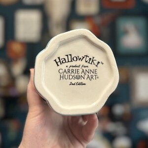 May include: A white ceramic mug base with the text "Hallowtiki" and "a product from CARRIE ANNE HUDSON ART" in black. The words "2nd Edition" are also printed on the base. The mug is held by a person.