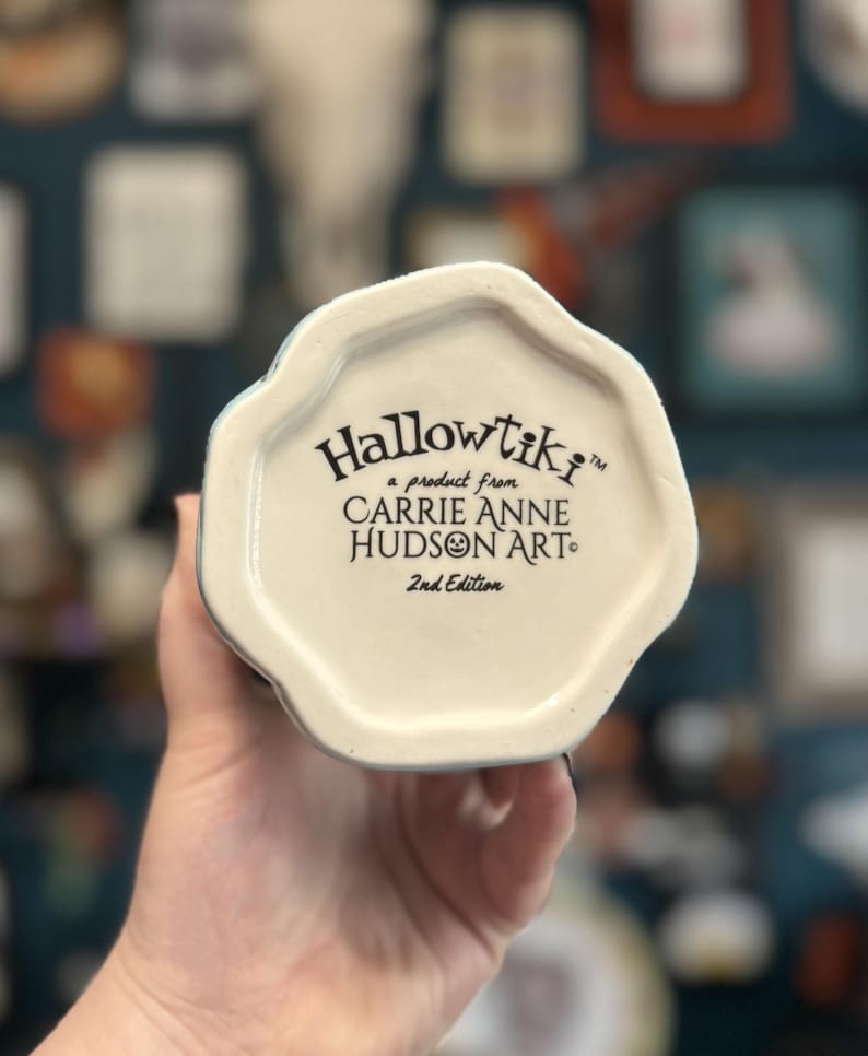 May include: A white ceramic mug base with the text "Hallowtiki" and "a product from CARRIE ANNE HUDSON ART" in black. The words "2nd Edition" are also printed on the base. The mug is held by a person.