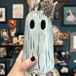 May include: A light blue ghost-shaped ceramic tumbler with black vertical lines and two large eye-shaped openings. The tumbler has a black and white striped straw and a small black and white paper umbrella. The tumbler is a Halloween-themed item.