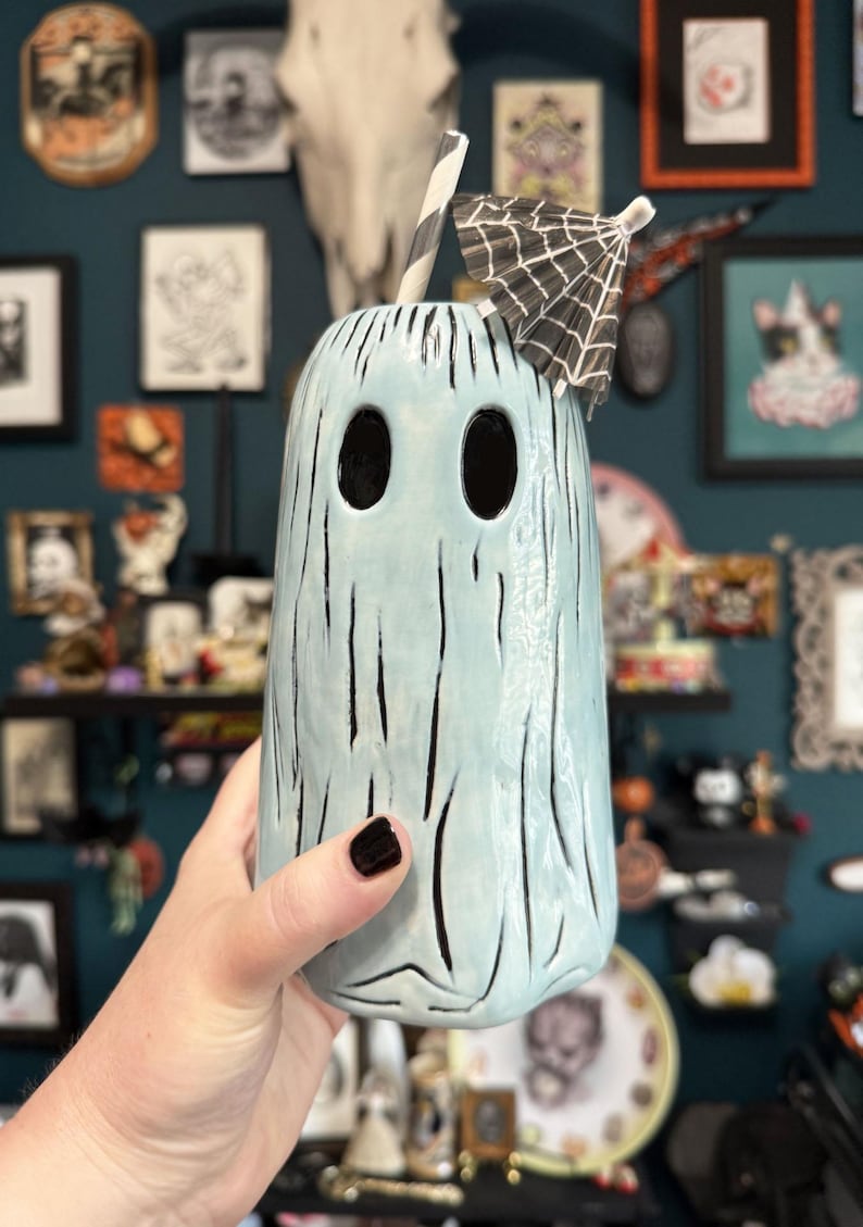 May include: A light blue ghost-shaped ceramic tumbler with black vertical lines and two large eye-shaped openings. The tumbler has a black and white striped straw and a small black and white paper umbrella. The tumbler is a Halloween-themed item.