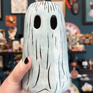 May include: A light blue ceramic ghost-shaped vase with black vertical lines and large oval eye holes. The vase is held by a hand with dark nail polish. The vase has a textured surface and is part of a Halloween or spooky-themed collection.