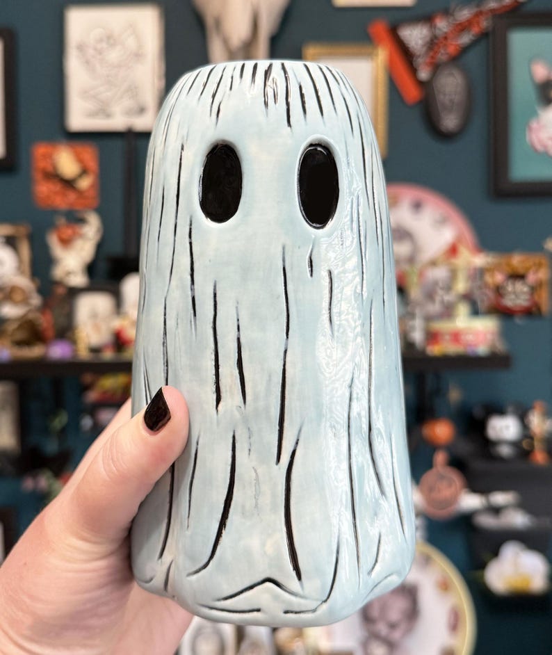 May include: A light blue ceramic ghost-shaped vase with black vertical lines and large oval eye holes. The vase is held by a hand with dark nail polish. The vase has a textured surface and is part of a Halloween or spooky-themed collection.
