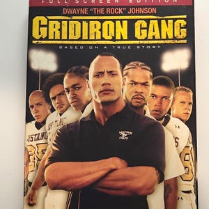 May include: A DVD case for the movie Gridiron Gang featuring Dwayne The Rock Johnson. The cover displays the title in yellow text, the phrase Based on a True Story, and a group of football players in white jerseys behind the main actor.
