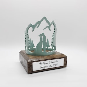 May include: A teal blue metal wedding cake topper with a silhouette of a couple kissing in front of a mountain range and pine trees. The topper is on a wooden base with a silver plaque that reads "Billy & Danielle August 28, 2021".