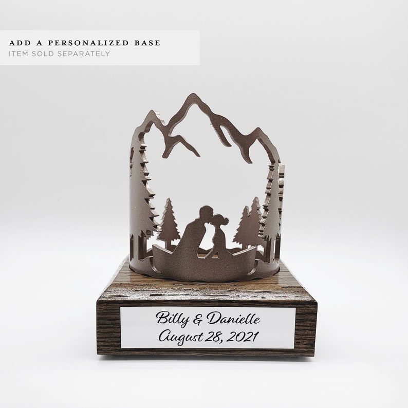 Op de afbeelding: Een houten bruidstaarttopper met een silhouet van een stel dat elkaar kust voor een bergketen en dennenbomen. De basis is gepersonaliseerd met de tekst "Billy & Danielle August 28, 2021".