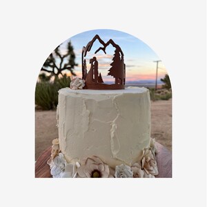 May include: A white wedding cake with buttercream frosting and a mountain range silhouette topper. The cake is decorated with paper flowers in shades of brown and white. The cake is set against a backdrop of a desert sunset.