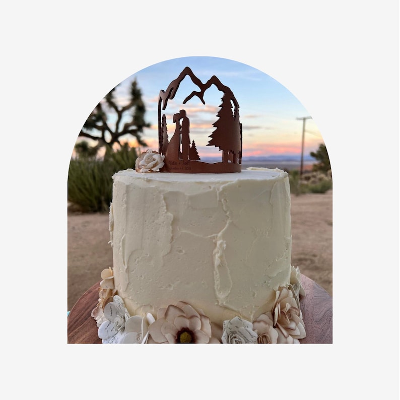May include: A white wedding cake with buttercream frosting and a mountain range silhouette topper. The cake is decorated with paper flowers in shades of brown and white. The cake is set against a backdrop of a desert sunset.