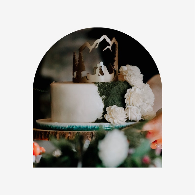Op de afbeelding: Een tweelaagse bruidstaart versierd met een groene mosachtige frosting en witte bloemen. De bovenste laag heeft een gouden bergketen topper met een silhouet van een stel.