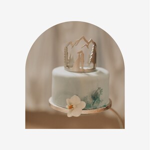 May include: A two-tiered wedding cake with a light blue frosting and a silver topper featuring a mountain range silhouette with a couple standing in front of it. The cake is decorated with a white orchid.