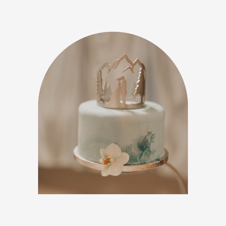 May include: A two-tiered wedding cake with a light blue frosting and a silver topper featuring a mountain range silhouette with a couple standing in front of it. The cake is decorated with a white orchid.