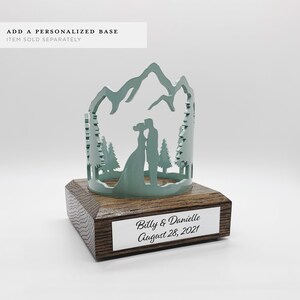 May include: A teal blue metal silhouette of a couple kissing in a mountain scene with trees. The silhouette is on a wooden base with a silver plaque that reads "Billy & Danielle August 28, 2021".