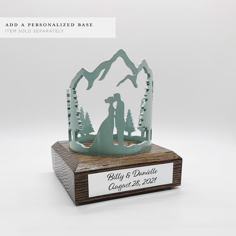 May include: A teal blue metal silhouette of a couple kissing in a mountain scene with trees. The silhouette is on a wooden base with a silver plaque that reads "Billy & Danielle August 28, 2021".