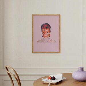 May include: A framed print of a portrait of David Bowie with a pink background. The portrait is in color and shows Bowie with his signature red lightning bolt makeup.