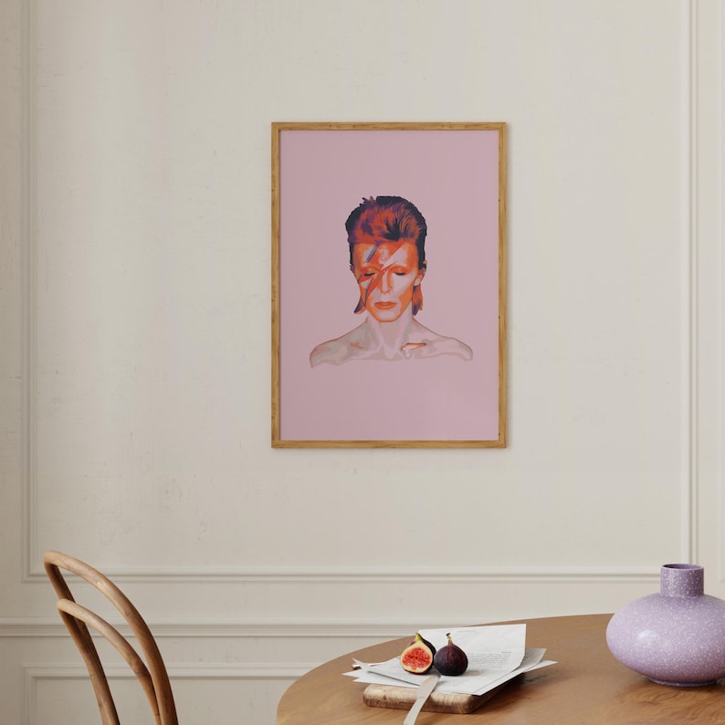 May include: A framed print of a portrait of David Bowie with a pink background. The portrait is in color and shows Bowie with his signature red lightning bolt makeup.