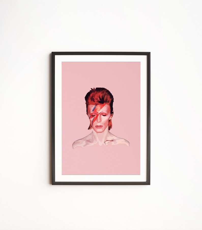 May include: A framed print of a portrait of David Bowie with a red and blue lightning bolt painted across his face. The background is a light pink color.