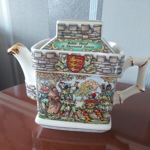 Peut inclure: Théière en céramique décorative avec un design en forme de château, représentant Robin des Bois dans la forêt de Sherwood. La théière a une base blanche avec des détails dorés sur la poignée et le bec. Le texte "Robin Hood in Sherwood Forest" est visible.