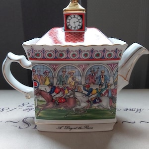 Puede incluir: Tetera "A Day at the Races". La tetera de porcelana blanca presenta una ilustración detallada de una carrera de caballos con espectadores. Una torre del reloj se encuentra en la tapa. La tetera tiene un asa y un pico dorados.