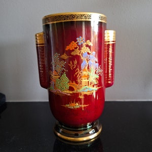 Puede incluir: Un jarrón de cerámica rojo con detalles dorados y una escena pintada de un jardín japonés. El jarrón tiene dos asas a los lados.