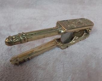 May include: A vintage brass nutcracker with a lever design. The nutcracker has a rectangular head with embossed details and a rounded handle. The brass has a warm, golden hue, and the tool appears to be well-used.