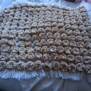 以下が含まれることがあります： 白地を背景に、ベージュのポンポンが格子状に配置された手作りのブランケット。ブランケットには白いフリンジの縁取りがあります。ポンポンは密に詰め込まれており、テクスチャのある表面を作り出しています。この居心地の良いブランケットは、暖かさとスタイルを加えるのに最適です。