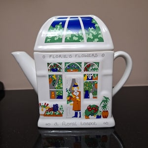 Puede incluir: Tetera de cerámica blanca con un diseño floral. La tetera tiene forma de una pequeña casa con un techo verde y ventanas. El texto "Florie's Flowers" y "a floral teapot" está impreso en la tetera.