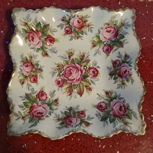 Op de afbeelding: Vierkante, crèmekleurige keramische schaal met een geschulpte rand, versierd met roze rozen en groene bladeren. De rand is afgewerkt met goud. Een decoratief stuk om te serveren of tentoon te stellen.