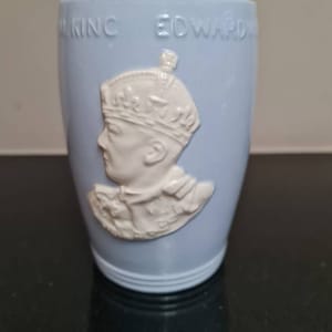 May include: A pale blue ceramic mug featuring the embossed text "KING EDWARD" and a white relief portrait of a crowned person. The mug is cylindrical in shape.