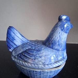 May include: A ceramic lidded container shaped like a hen sitting on a woven basket base. The piece features a blue and white transferware pattern with detailed feather textures on the hen and a cross-hatched basket weave design on the lower section.