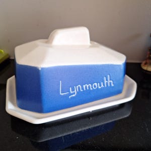 Puede incluir: Una mantequera de cerámica blanca y azul con la palabra "Lynmouth" en escritura blanca. El plato tiene una base rectangular y una tapa con asa. La sección azul es de color liso.