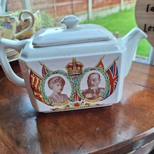 May include: White ceramic teapot with a lid, handle, and spout. The teapot features portraits of Queen Mary and King George, with flags and text. The teapot is on a wooden surface.