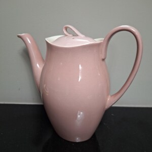 Puede incluir: Una tetera de cerámica de color rosa pálido con tapa y asa blancas. La tetera tiene un cuerpo redondeado y un pico en el lateral.