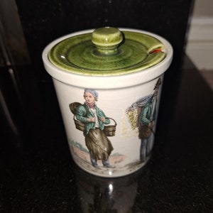 May include: A white ceramic jar with a green lid and a painted scene of two people carrying baskets. The jar has a small spout on the lid.