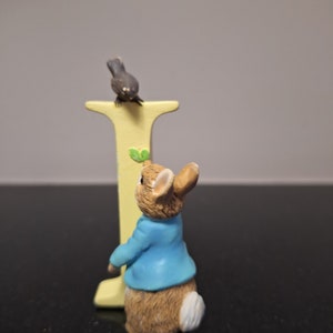 May include: A yellow letter I with a blue-clad rabbit figurine standing in front of it. A small brown bird is perched on top of the letter.