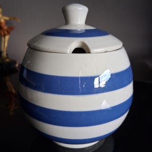 Puede incluir: Un azucarero de cerámica a rayas azules y blancas, con tapa y ranura para cuchara. El cuenco redondo se asienta sobre una pequeña base. La tapa tiene un pequeño asa. Un artículo de cocina clásico.