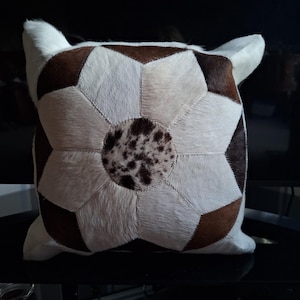 May include: A white and brown cowhide pillow with a starburst design. The pillow is made of different pieces of cowhide sewn together to create a geometric pattern.