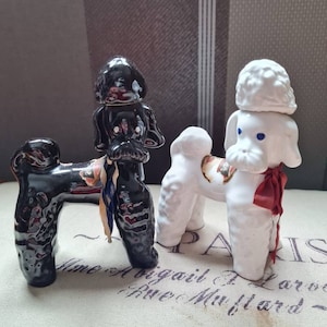 May include: Two ceramic poodle figurines, one black and one white, with red and blue ribbon accents. The figurines are standing on a white fabric surface with text.