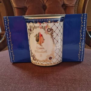 Peut inclure: Une tasse en céramique blanche et dorée avec un bord bleu et une boîte bleue. La tasse présente une image de la reine Elizabeth II et des accents dorés décoratifs. La tasse est présentée dans une boîte bleue avec des détails dorés.