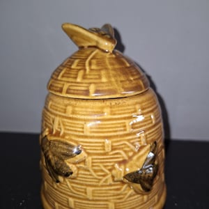 Puede incluir: Un tarro de miel de cerámica con forma de colmena con una abeja en la parte superior. El tarro es marrón con una superficie texturizada y tiene tres abejas en los lados.